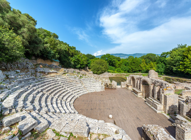 An ancient stone amphitheater with semi-circular seating surrounds a wooden stage, nestled in a lush, green forest. Ruins and arches are visible, under a bright blue sky.